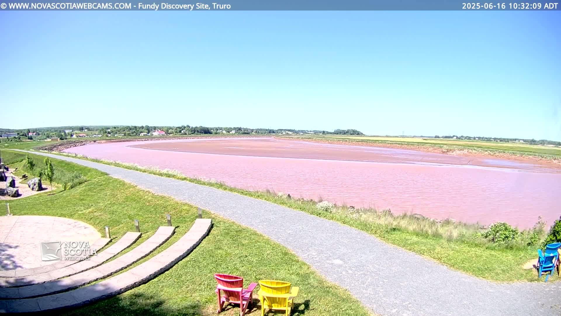 A pink-hued river or bay is seen from a grassy park with a paved path and colorful Adirondack chairs under a clear blue sky.