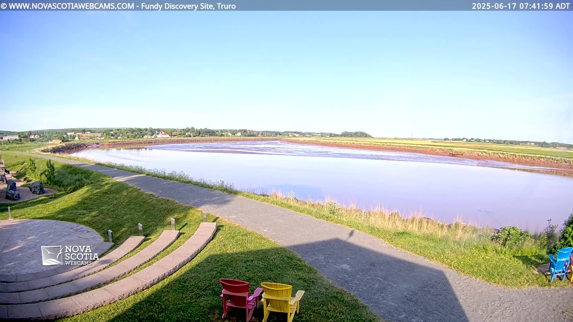 A calm body of water, under a clear blue sky, stretches out from a grassy area with a paved walkway and several colorful chairs.
