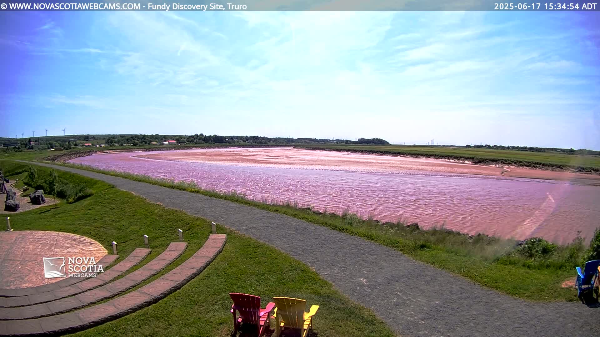 A pink-hued river flows through a grassy landscape under a mostly sunny sky, with a paved pathway and outdoor seating visible in the foreground.