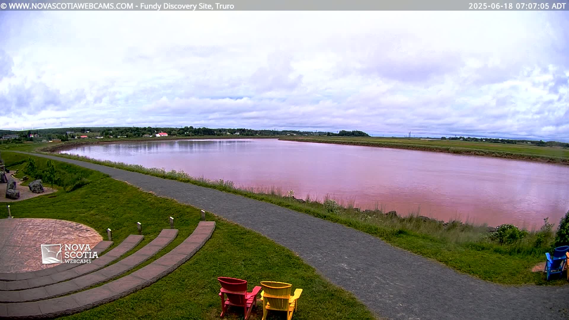 A wide, pinkish-red river flows under a partly cloudy sky, beside a paved walkway and grassy area with several colorful Adirondack chairs.