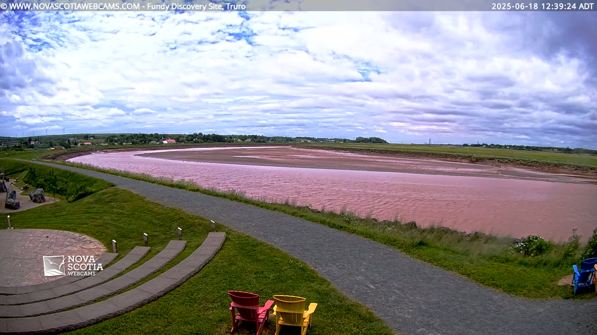 A pink river flows through a grassy landscape under a partly cloudy sky, with a paved pathway and several empty chairs visible in the foreground.