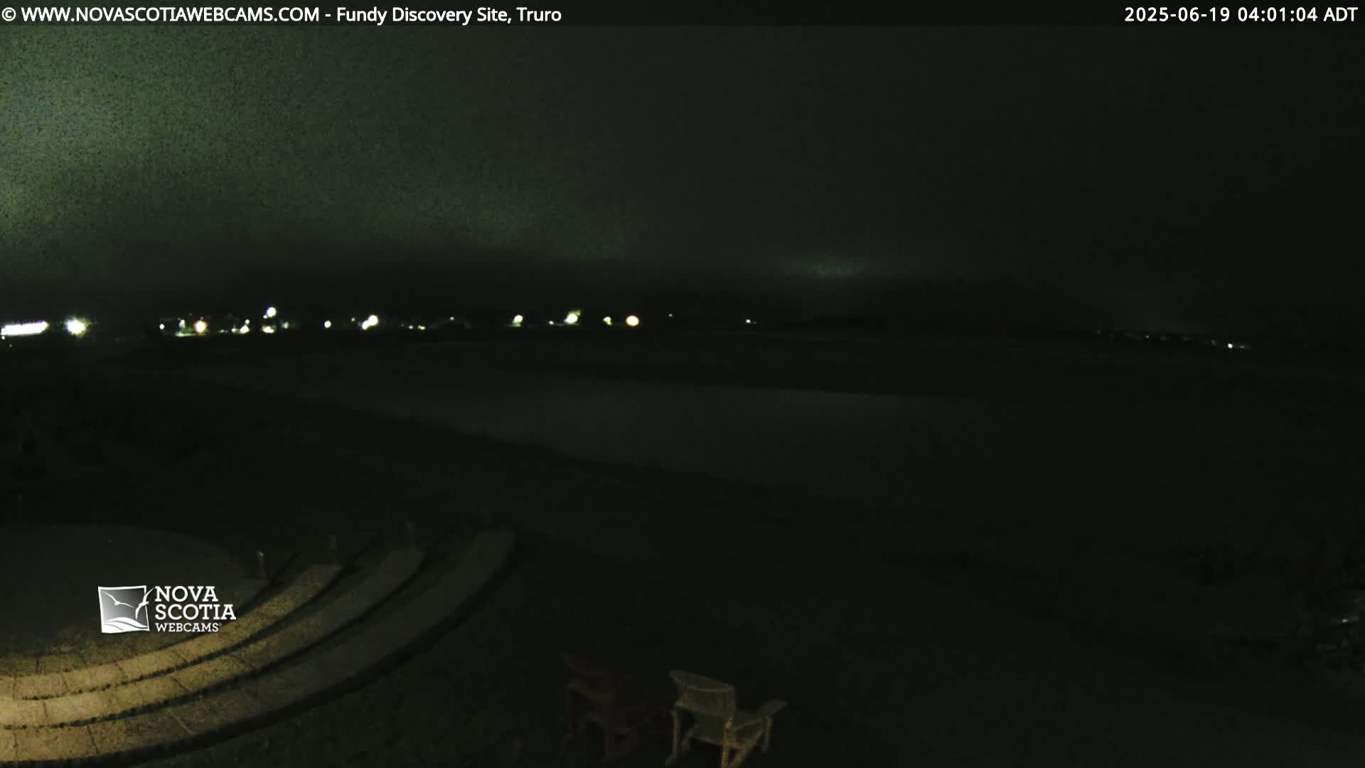 A dark, nighttime scene shows a mostly empty amphitheater overlooking a distant, dimly lit shoreline under a cloudy sky.