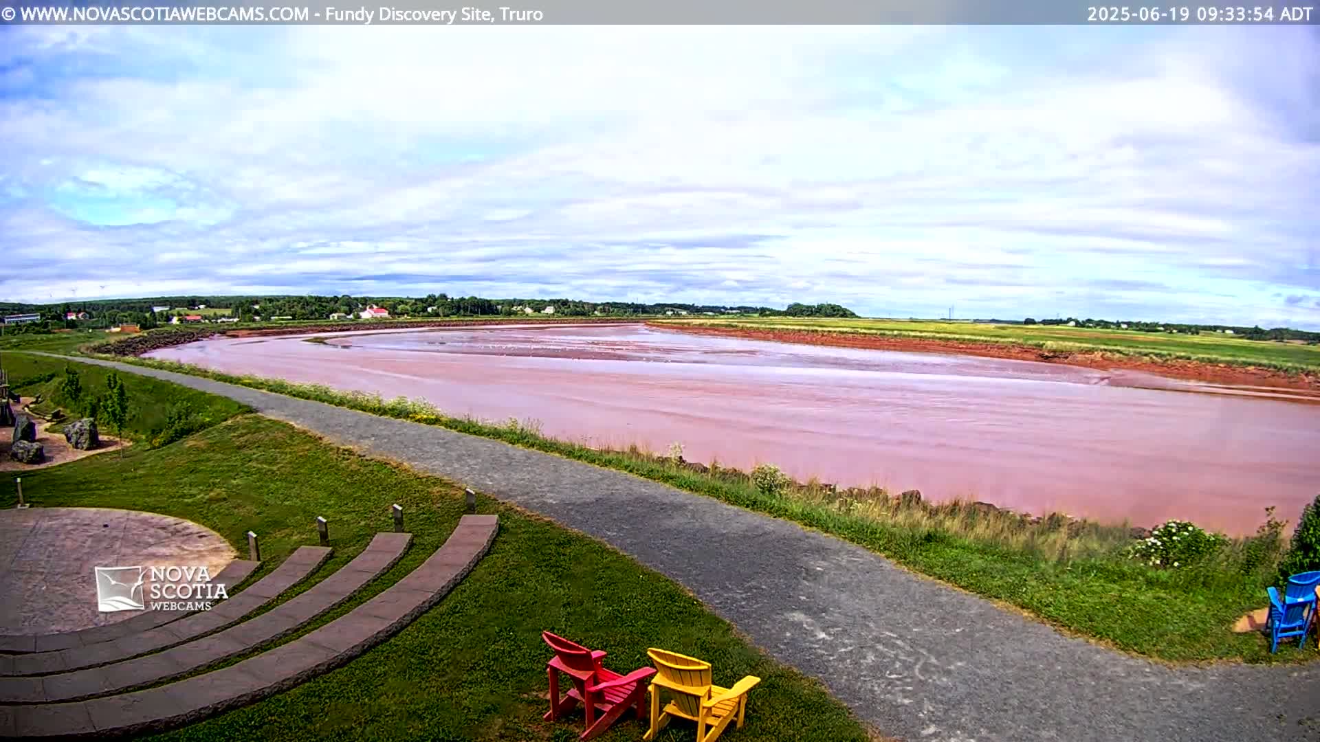 A pink-hued river flows through a grassy landscape under a partly cloudy sky, with a paved pathway and several Adirondack chairs in the foreground.