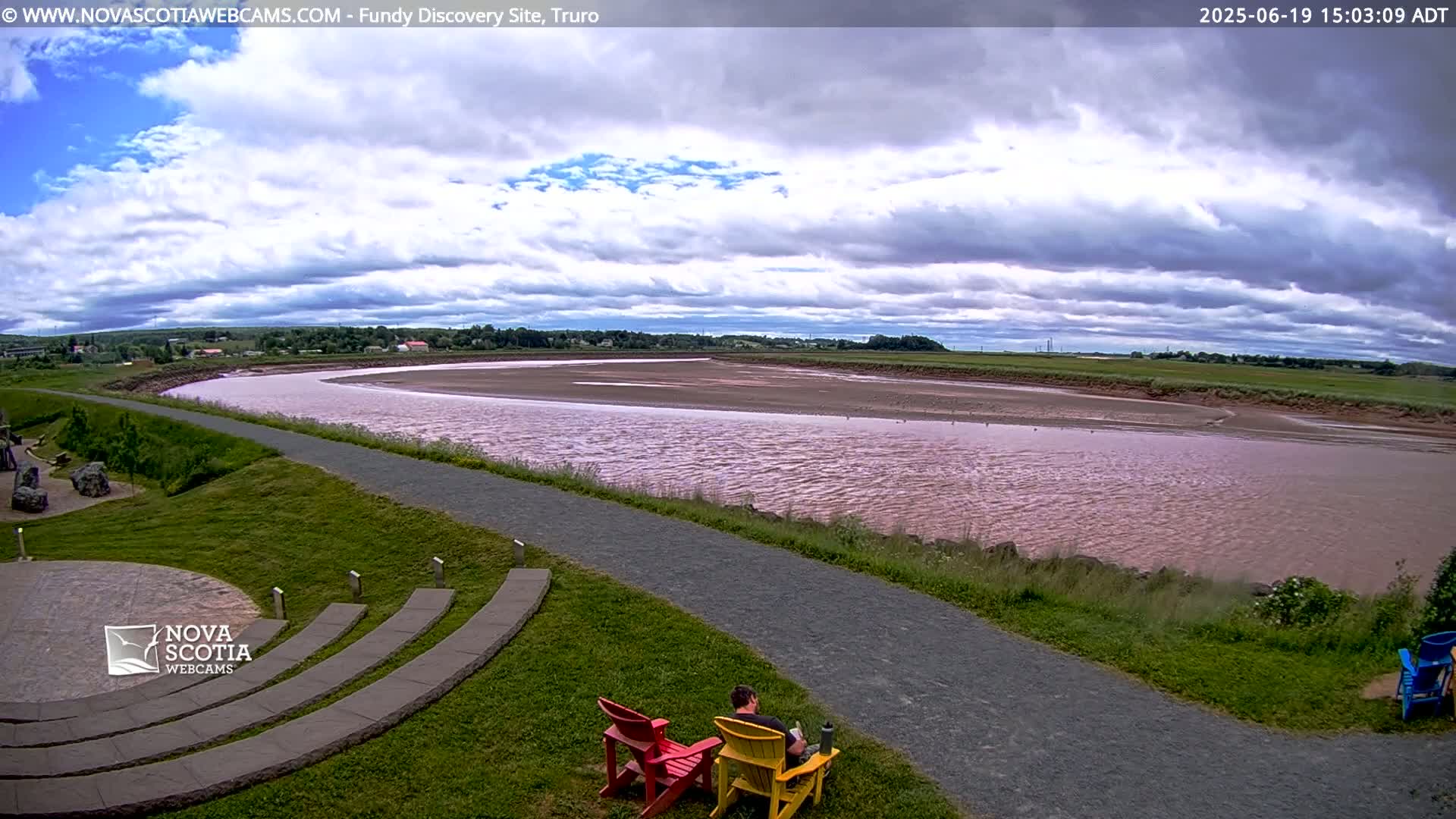 A man sits in a yellow lawn chair beside a red one overlooking a wide, pinkish-red river under a partly cloudy sky.