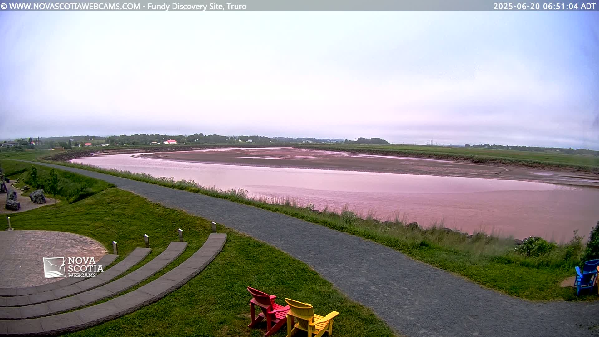 A wide, pink-hued river flows through a flat landscape under a cloudy sky, with a paved pathway and colorful chairs situated on a grassy area overlooking the water.