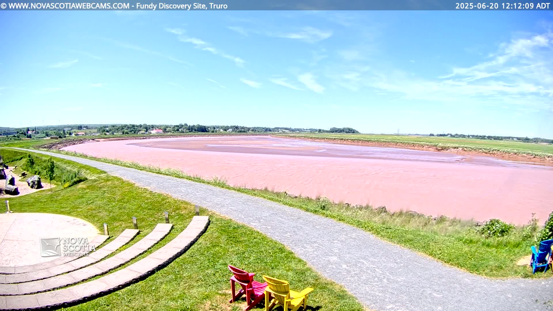 A pink-hued tidal river stretches into the distance under a mostly sunny sky, with a paved path and grassy area in the foreground, featuring a few colorful chairs.