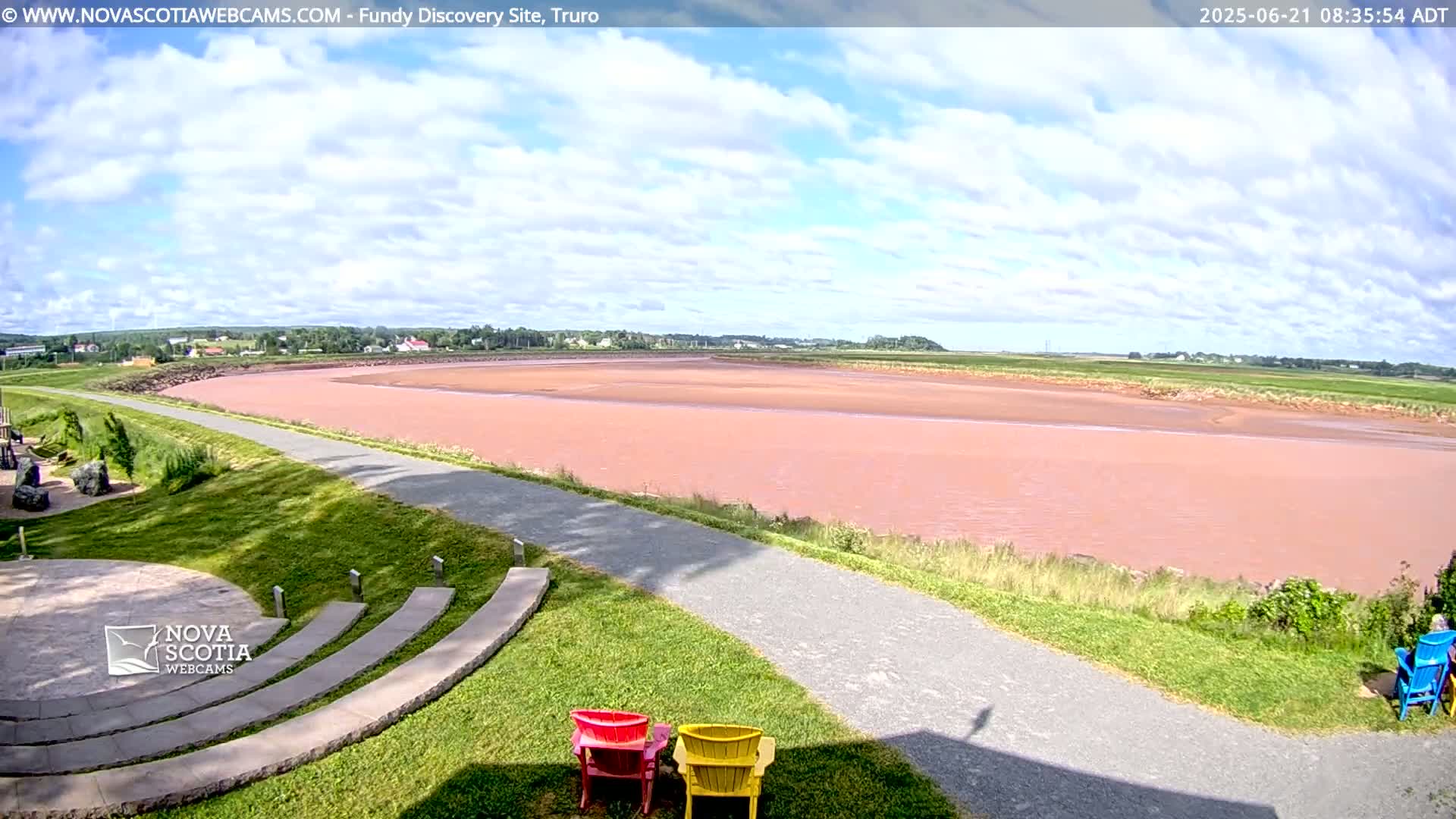 A wide, pinkish-red river flows through a flat landscape under a partly cloudy sky, with a paved pathway leading to an amphitheater-like seating area and a few colorful chairs.