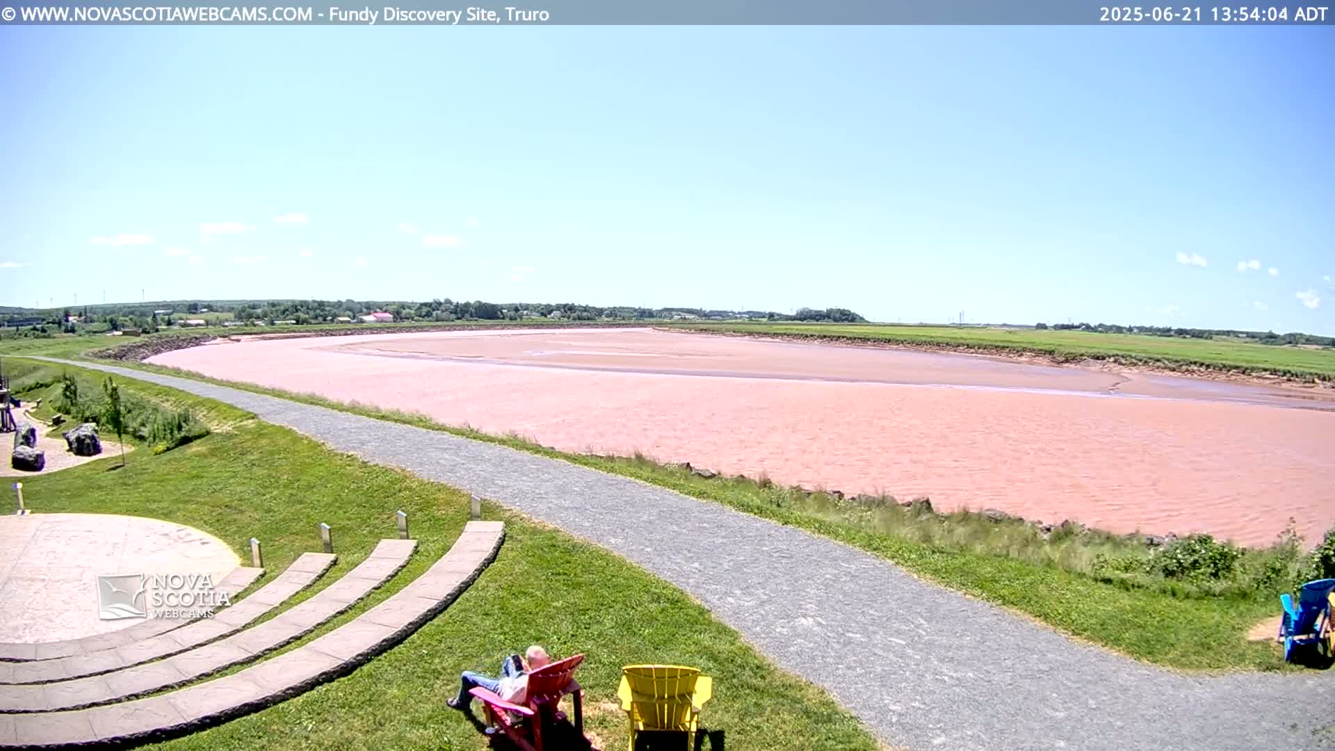 A pinkish-red river stretches across a grassy landscape under a clear blue sky, with two people relaxing in lawn chairs near a paved pathway.