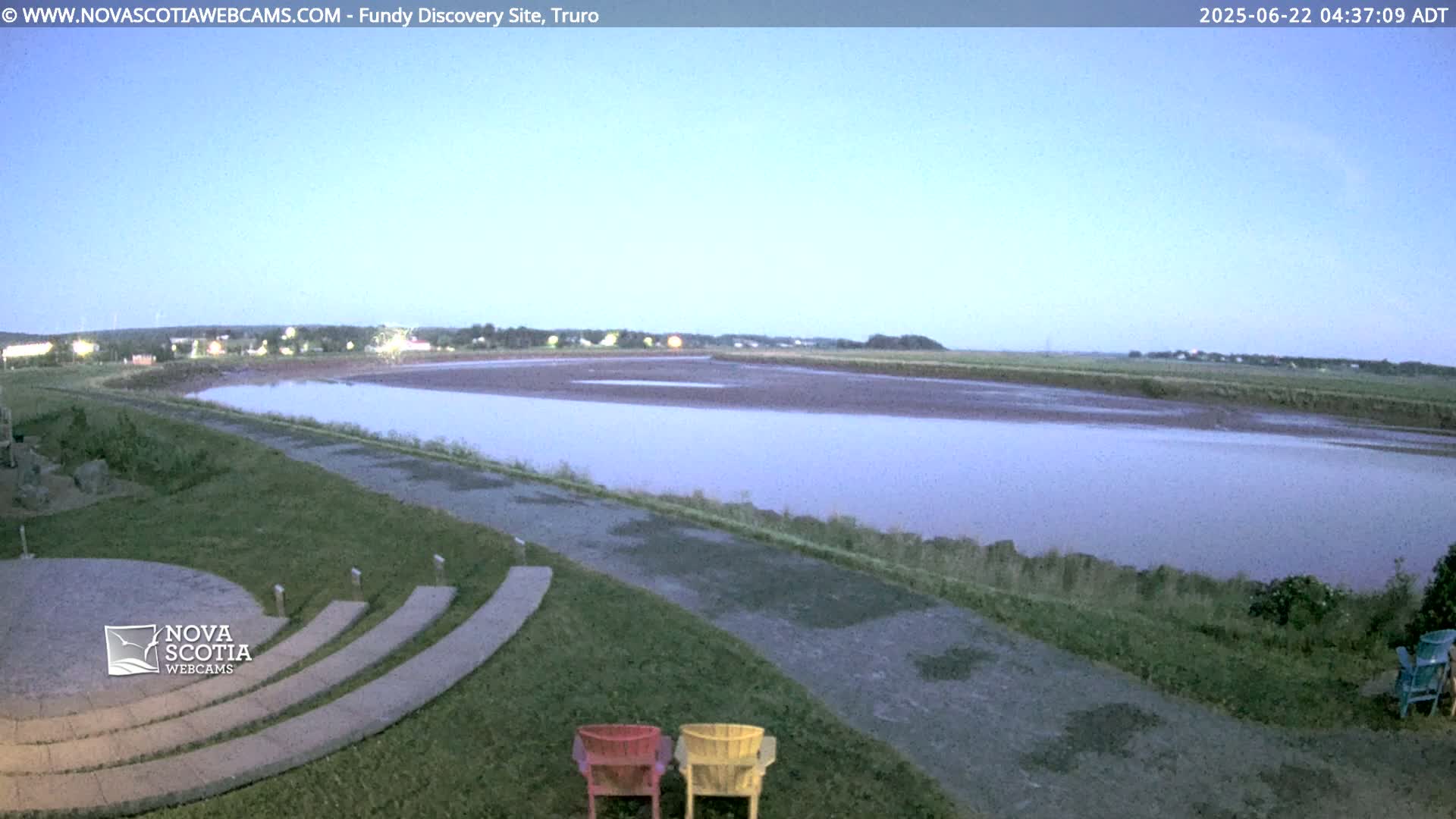 A calm, pale-blue-hued body of water stretches across a flat landscape under a clear twilight sky, with a paved walkway and a few empty chairs in the foreground.