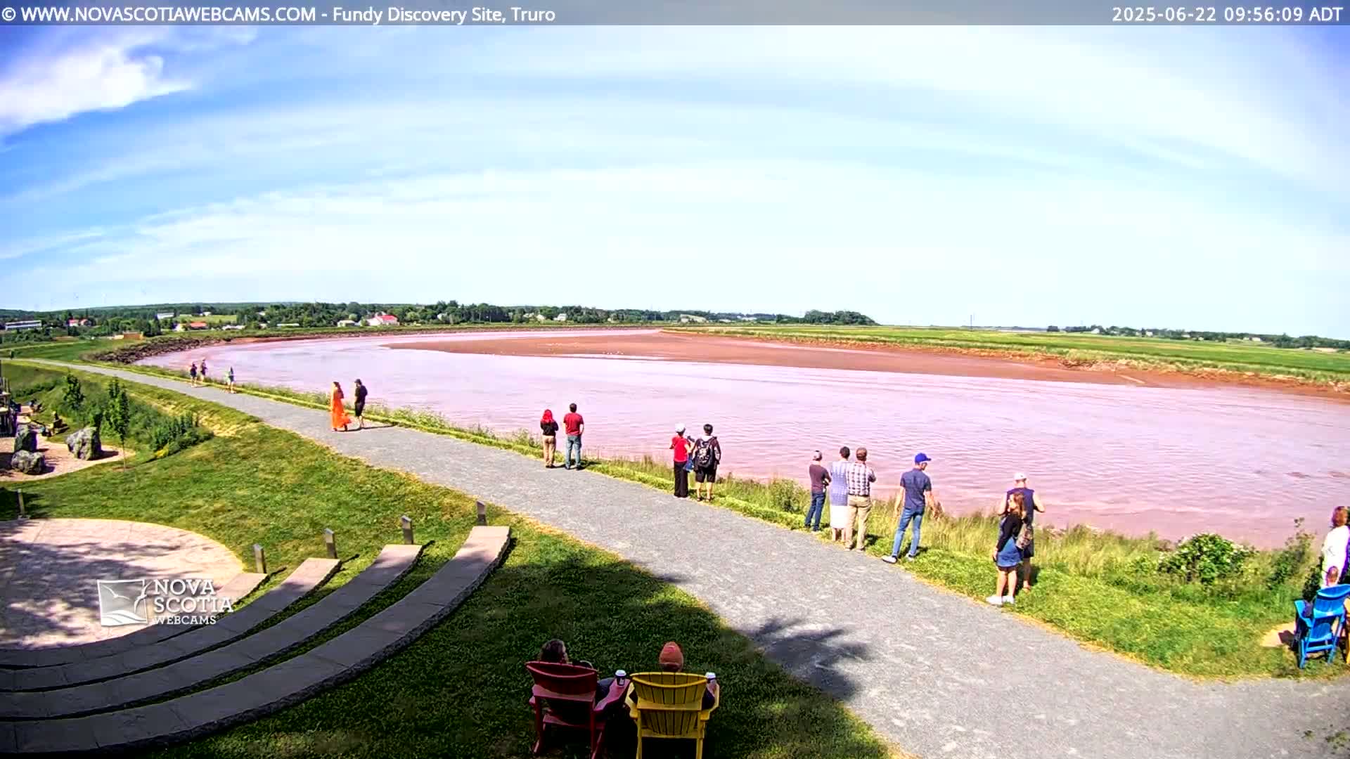 A wide, pinkish-red river flows under a mostly sunny sky, with several people standing along a paved path overlooking it, and two people seated in chairs nearby.