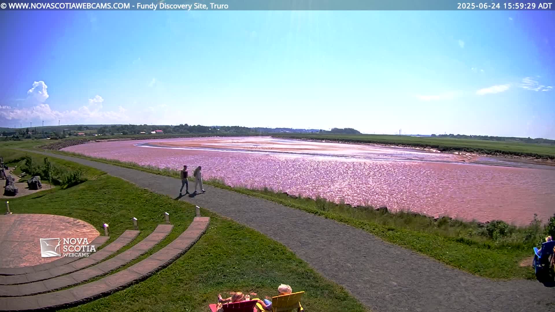 A pink-hued river flows past a paved walkway and grassy area with a few people walking and sitting, under a mostly sunny sky with some scattered clouds.