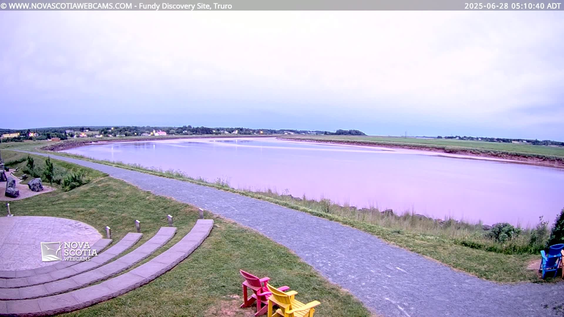 A calm, pinkish-purple river is seen from an elevated viewpoint, with a paved pathway leading to a stone amphitheater and a few colorful chairs in the foreground under a light cloudy sky.