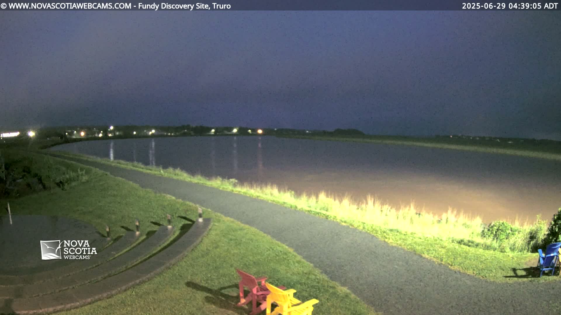 A calm, dark body of water is seen at twilight, next to a paved path and grassy area with several empty chairs.