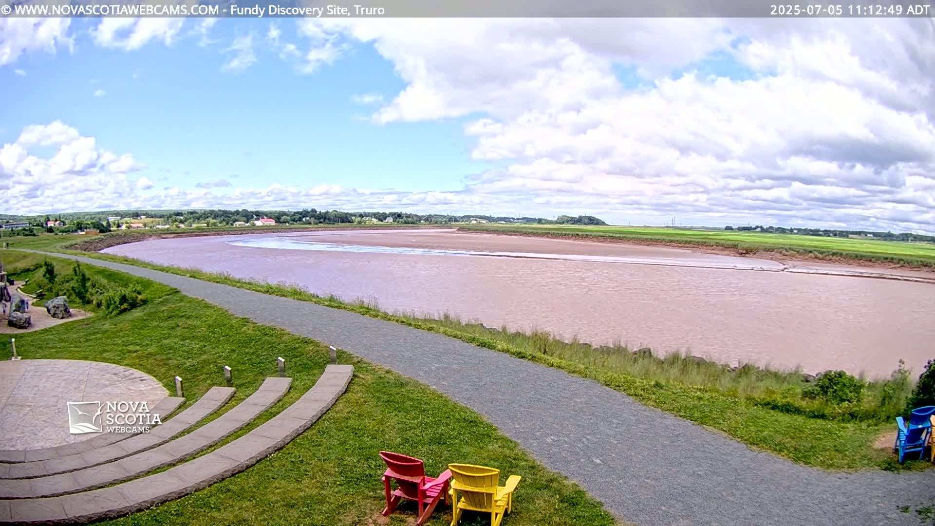 A wide, calm, pinkish-brown river flows through a green landscape under a partly cloudy sky, with a paved pathway and colorful chairs situated near the riverbank.