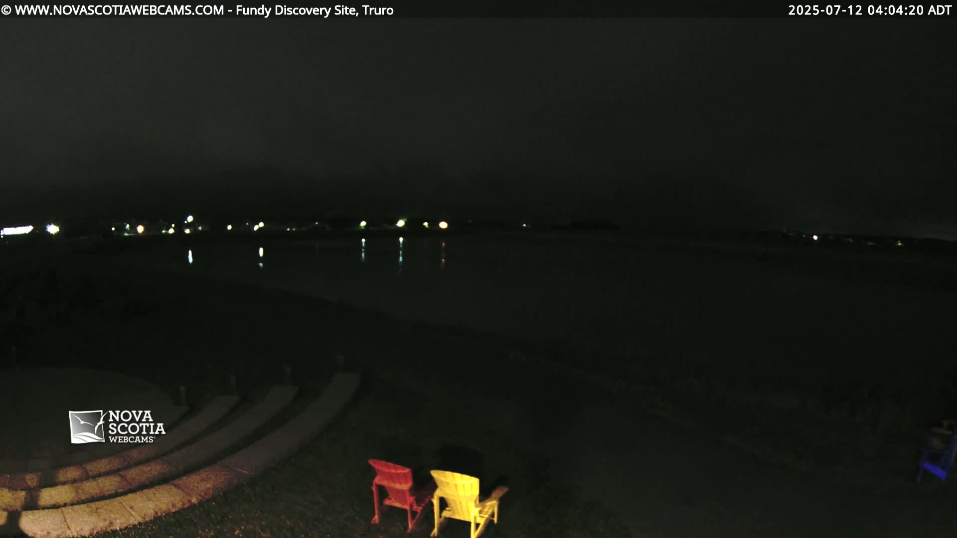 At nighttime, two Adirondack chairs sit on a grassy area overlooking a dark body of water reflecting distant lights under a mostly clear sky.