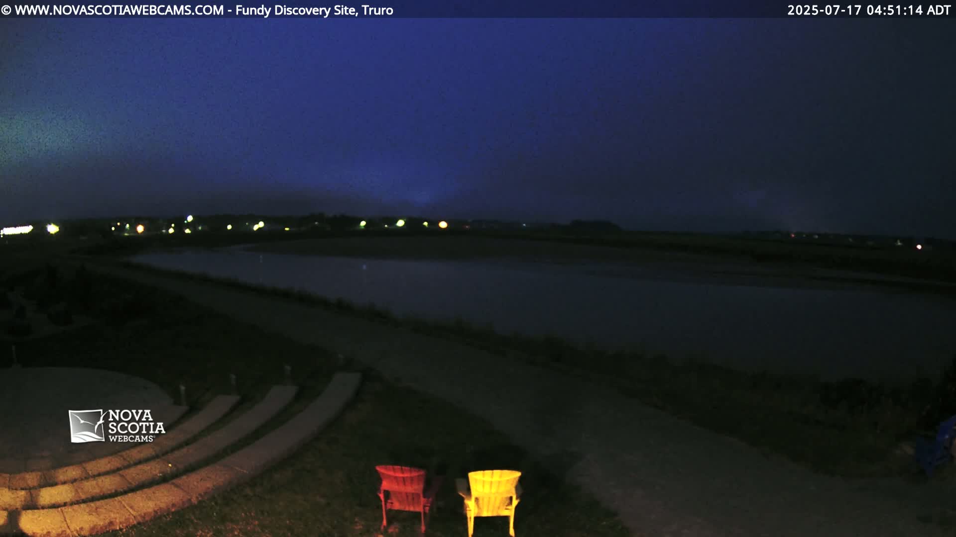 A dark, nighttime view of a calm body of water, a stone amphitheater, and two Adirondack chairs, under a twilight sky.