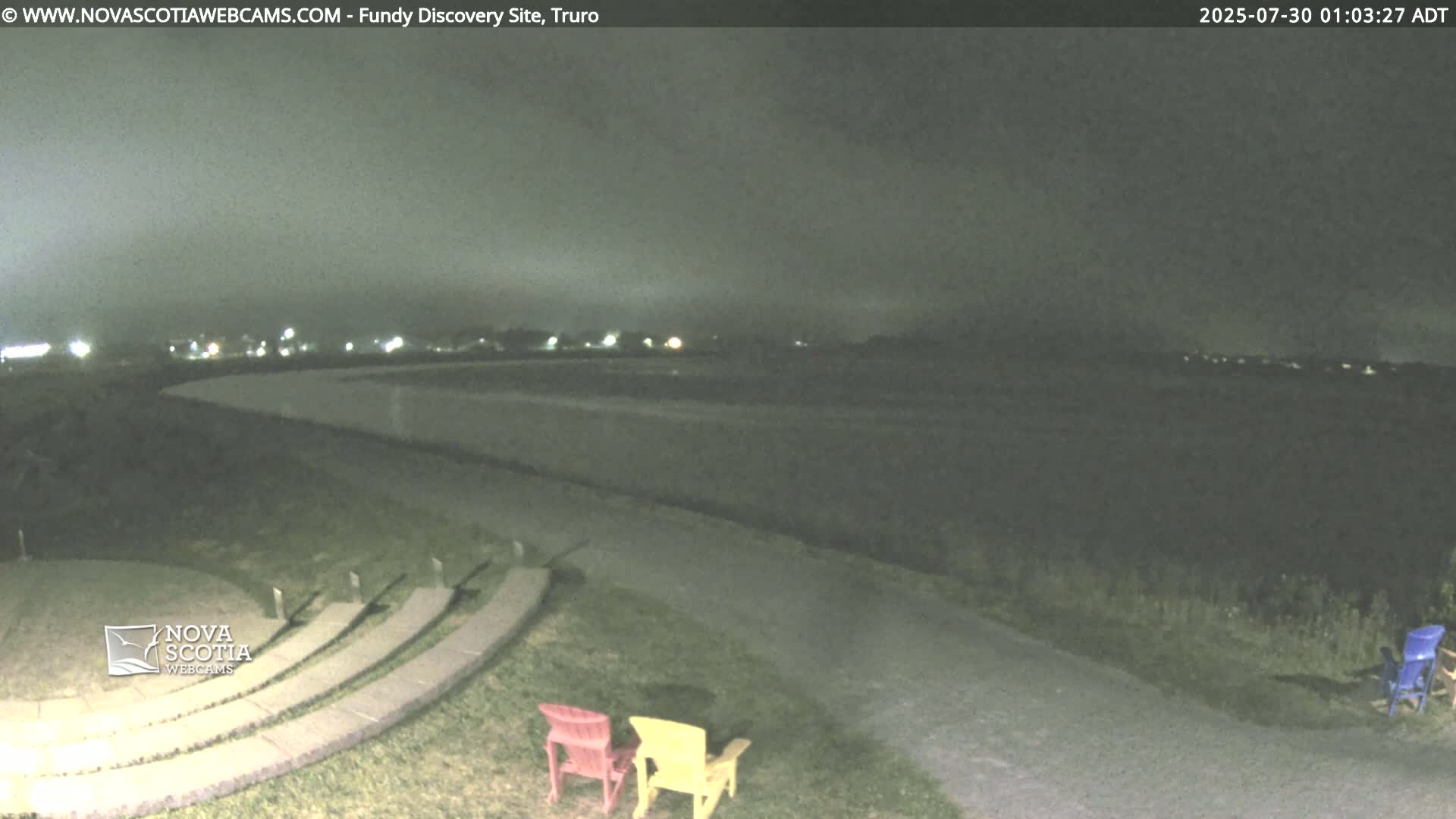 A dark, nighttime view of a calm body of water, a stone amphitheater, and two Adirondack chairs, under a twilight sky.