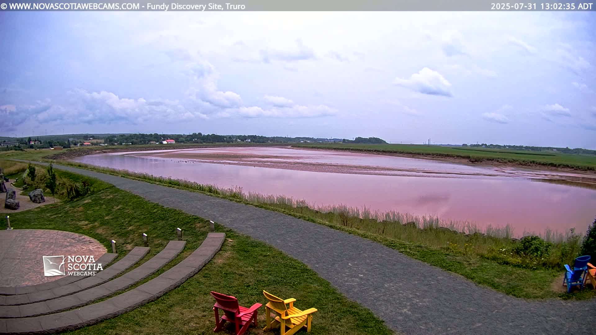 A wide, pink-tinged river is visible under a partly cloudy sky, with a paved pathway and grassy area in the foreground, featuring a few colorful Adirondack chairs.