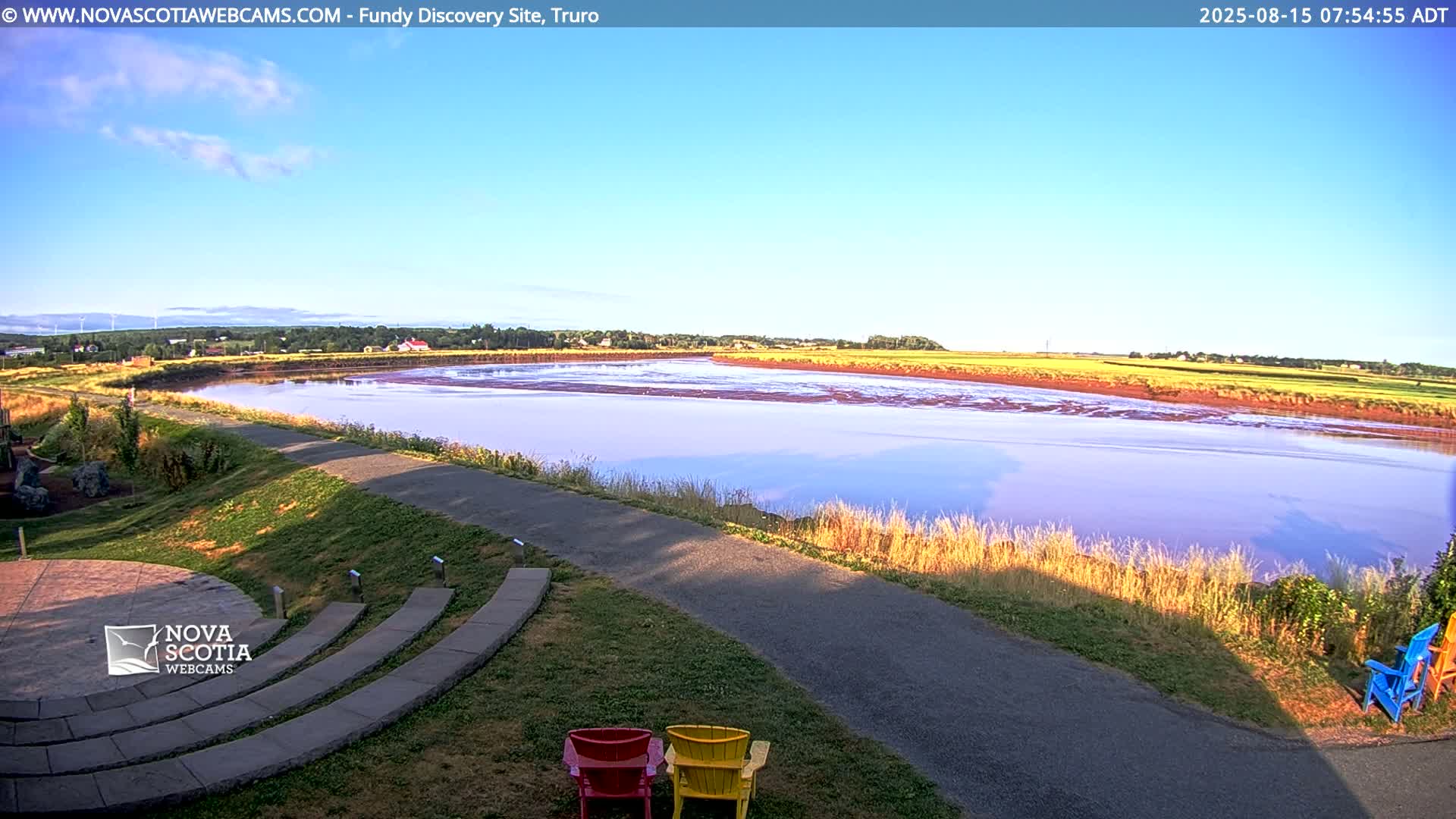 A wide, pink-tinged river is visible under a partly cloudy sky, with a paved pathway and grassy area in the foreground, featuring a few colorful Adirondack chairs.