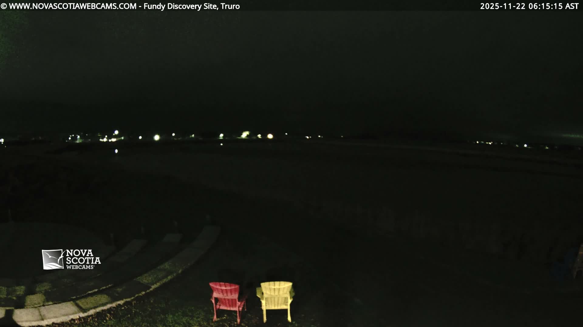 Two brightly colored Adirondack chairs sit in the foreground of a dark, overcast outdoor scene at night, with a distant array of blurred lights illuminating the horizon.
