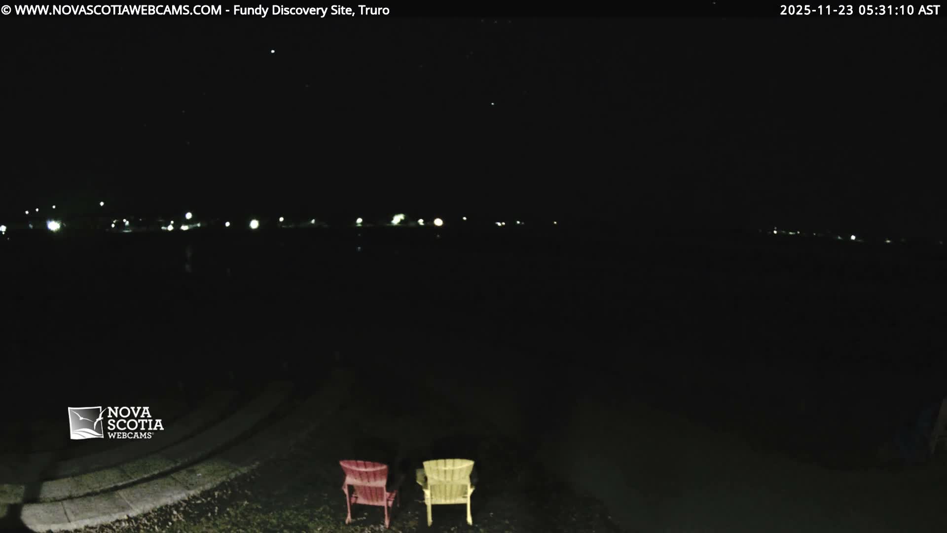 It is a dark and clear night, with a red and a yellow Adirondack chair visible on a grassy area in the foreground, facing distant lights across a body of water or open field.