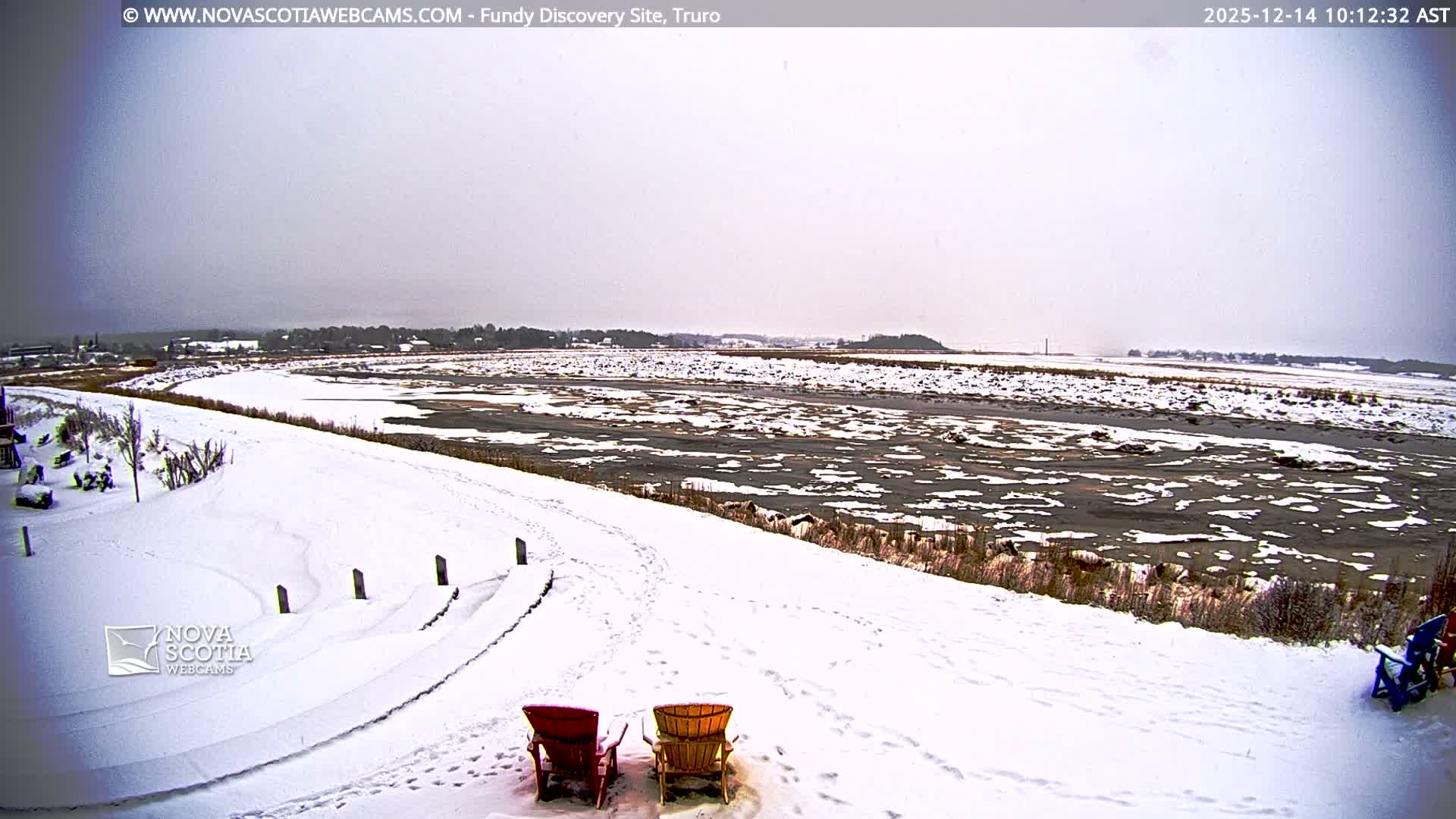 A snowy outdoor landscape at night depicts a path running alongside a body of water that reflects distant town lights, with tiered seating and two colorful Adirondack chairs prominent in the snow-covered foreground under a dark, overcast sky.