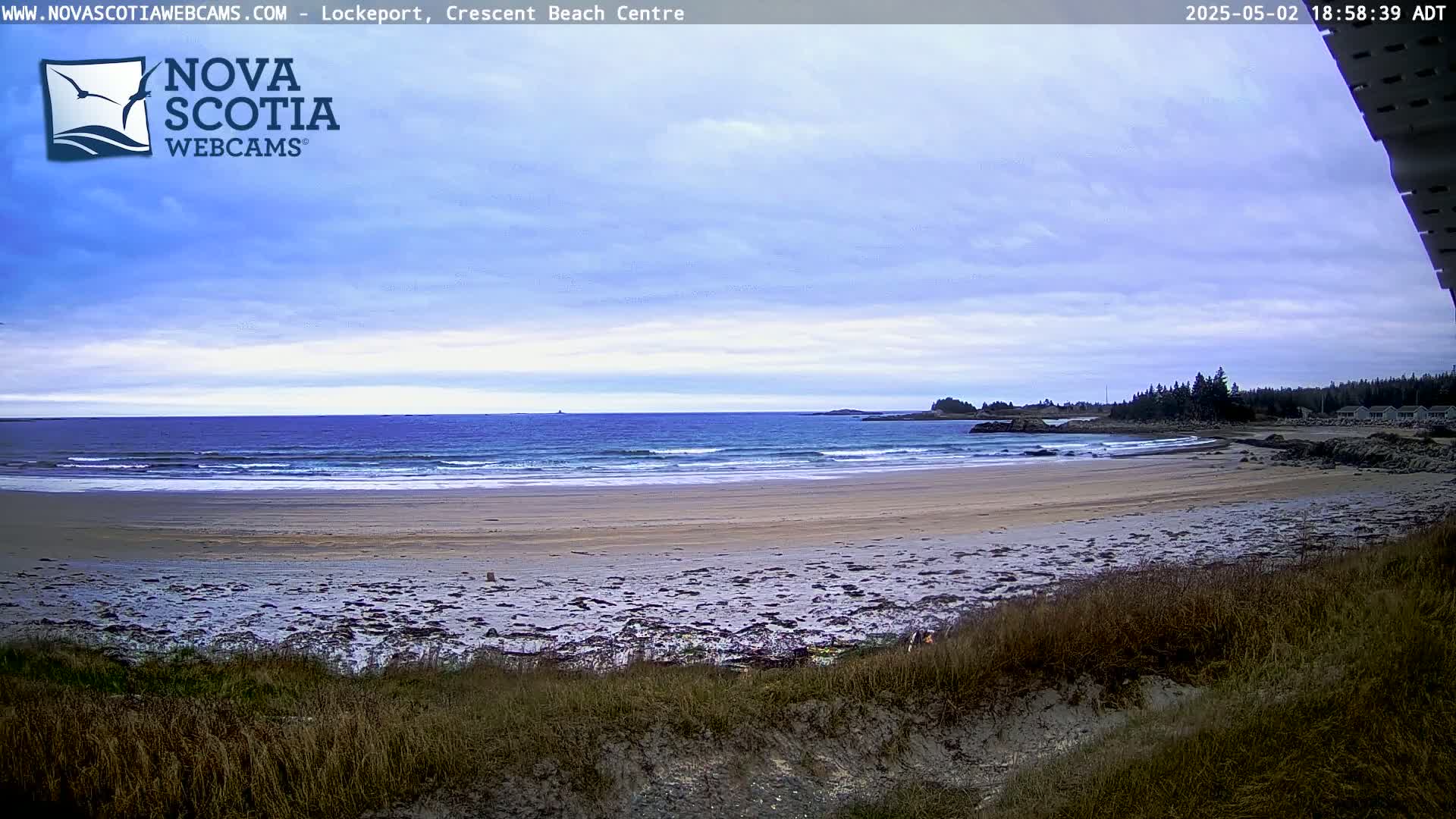 A sandy beach with gentle waves under a partly cloudy sky meets a calm ocean with a distant shoreline.