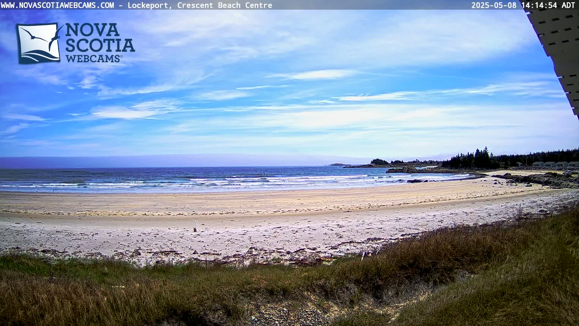 A sandy beach with gentle waves under a mostly sunny sky stretches to a coastline with dark green trees.