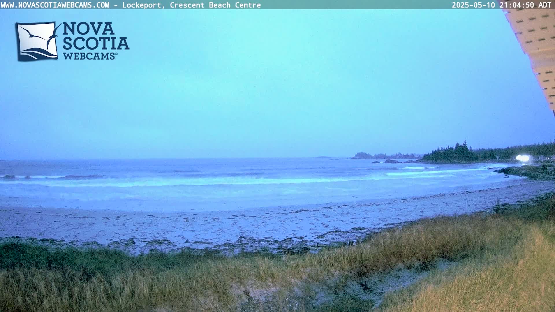 A mostly cloudy, twilight scene shows a sandy beach with small waves lapping the shore, backed by a line of trees.