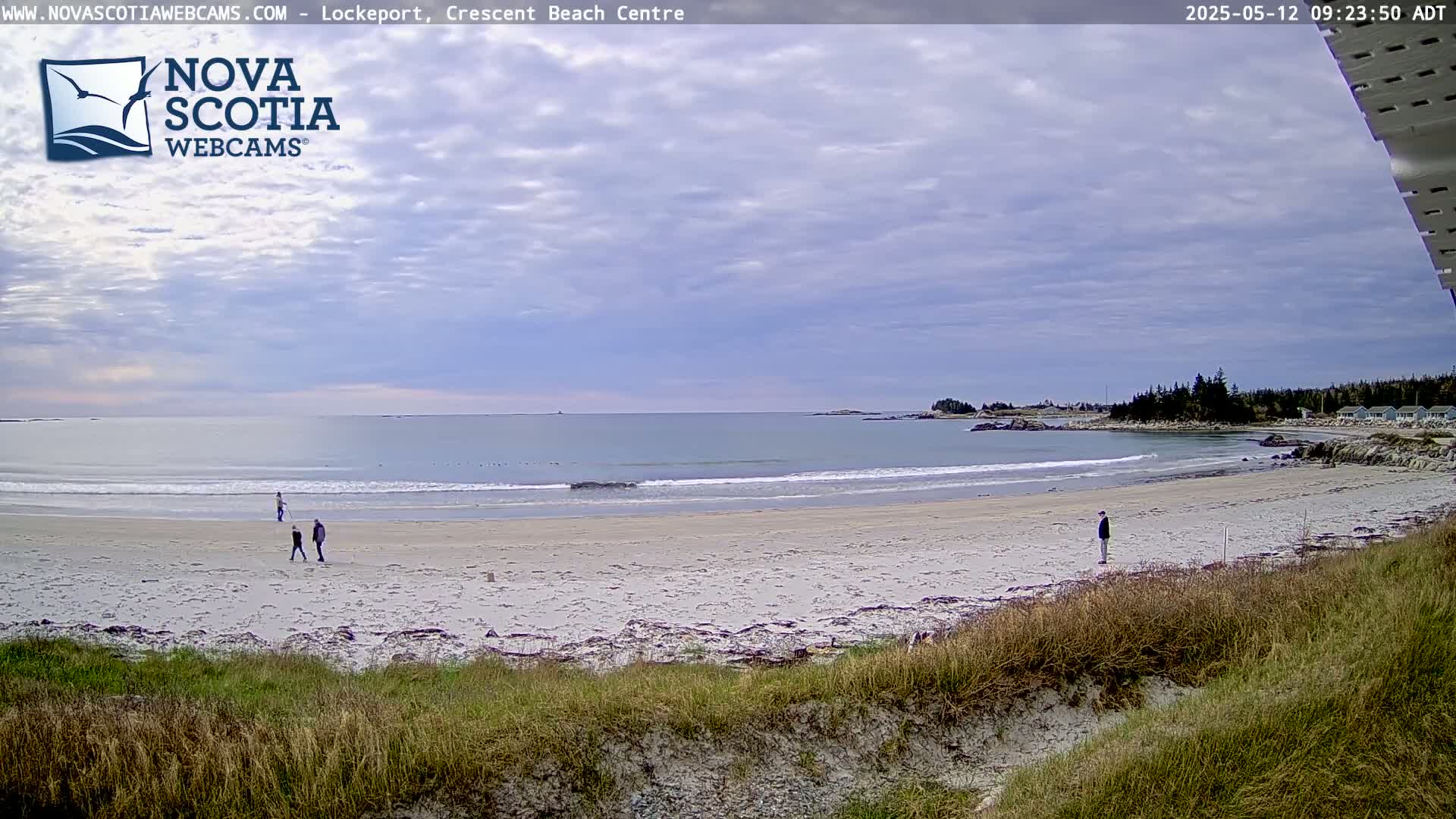 A sandy beach with a few people walking on it, under a partly cloudy sky, next to calm ocean waves.