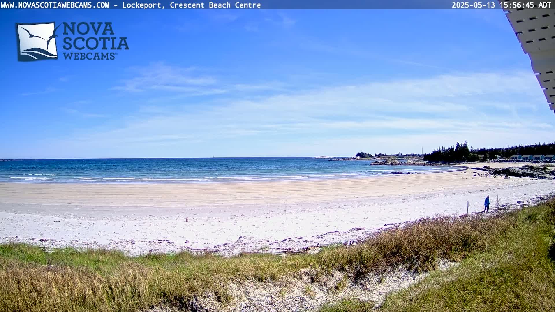 A sandy beach stretches to a calm, blue ocean under a mostly sunny sky, with a single person visible walking near the water's edge.