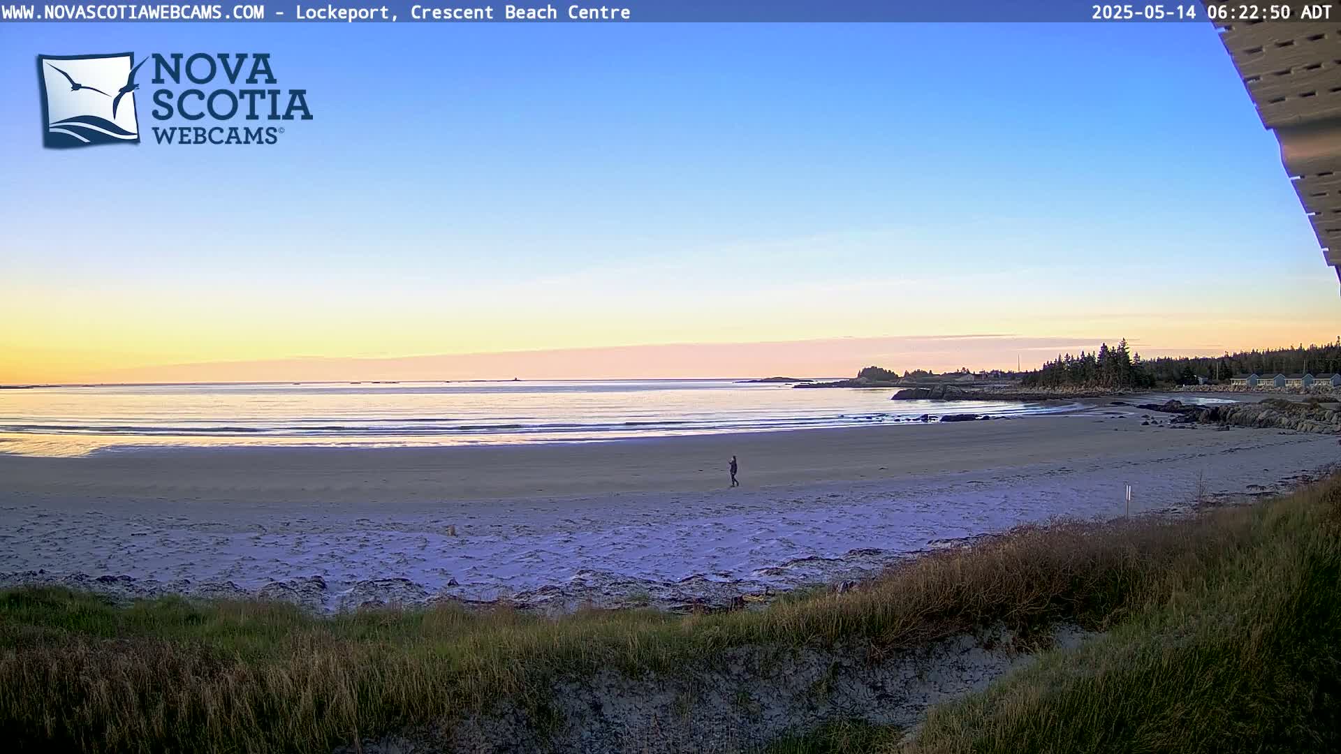 A person walks along a sandy beach at sunset, with calm ocean waves lapping the shore and a quiet coastline in the distance.