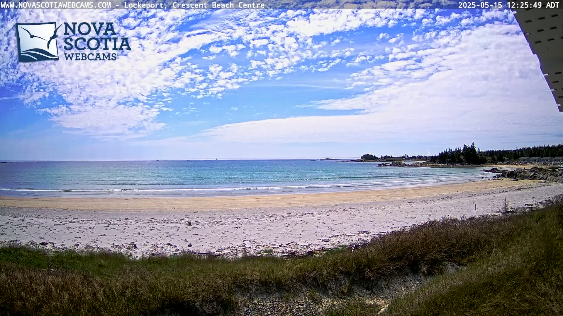 A sandy beach with calm, light-blue water stretches to a distant shoreline under a partly cloudy blue sky.