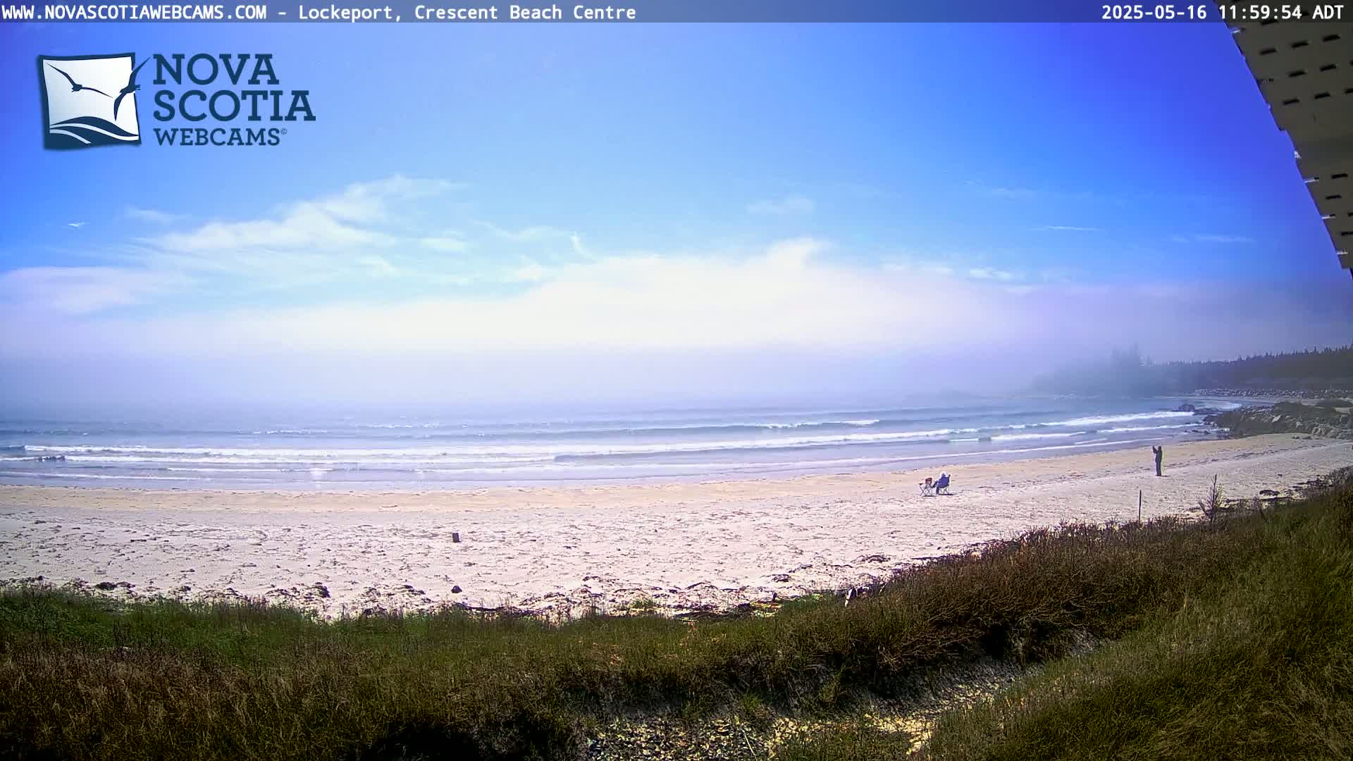 A sandy beach with a few people is visible, with small waves rolling onto the shore under a mostly sunny sky with some light cloud cover and a slight haze on the horizon.