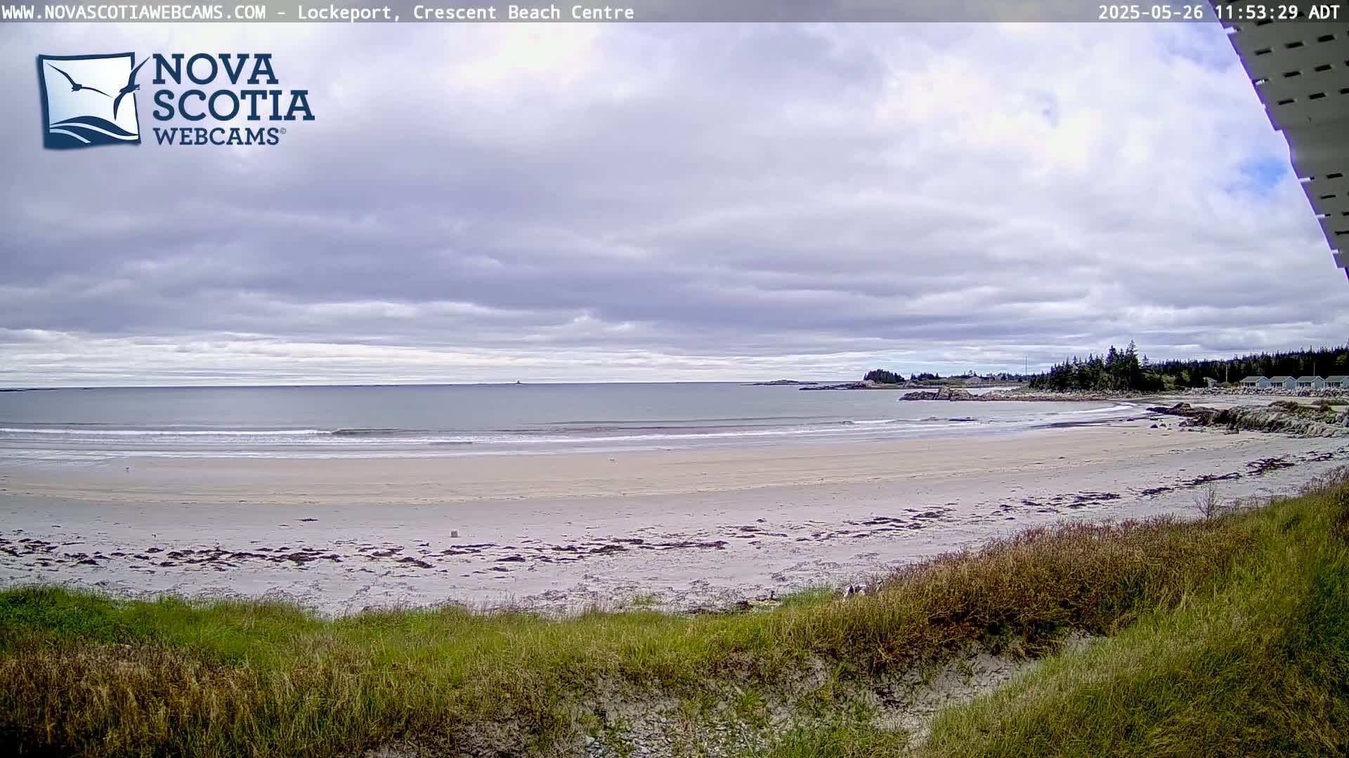 A sandy beach with small waves on a partly cloudy day is viewed from a grassy dune.