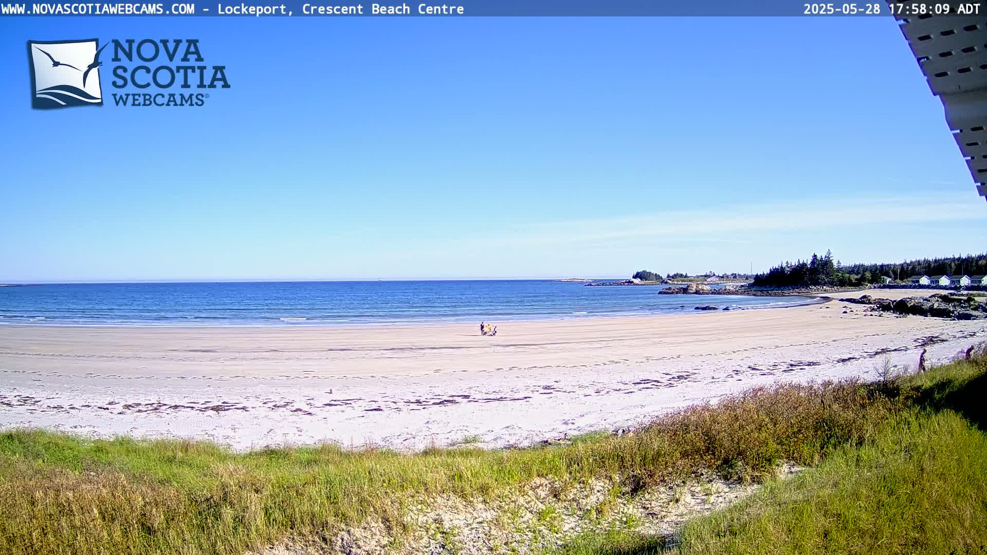 A sandy beach with calm, blue water under a clear blue sky, a few people are visible near the water's edge.