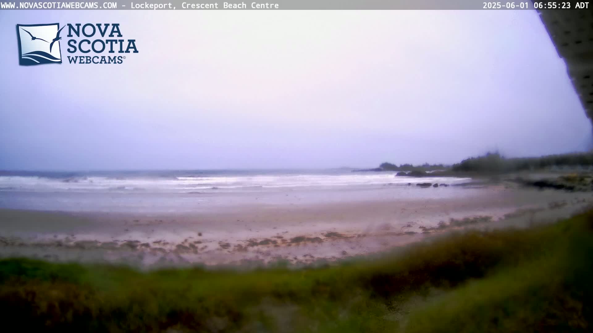 A mostly cloudy day shows a wide sandy beach with small waves lapping the shore, and a rocky outcrop visible in the distance.