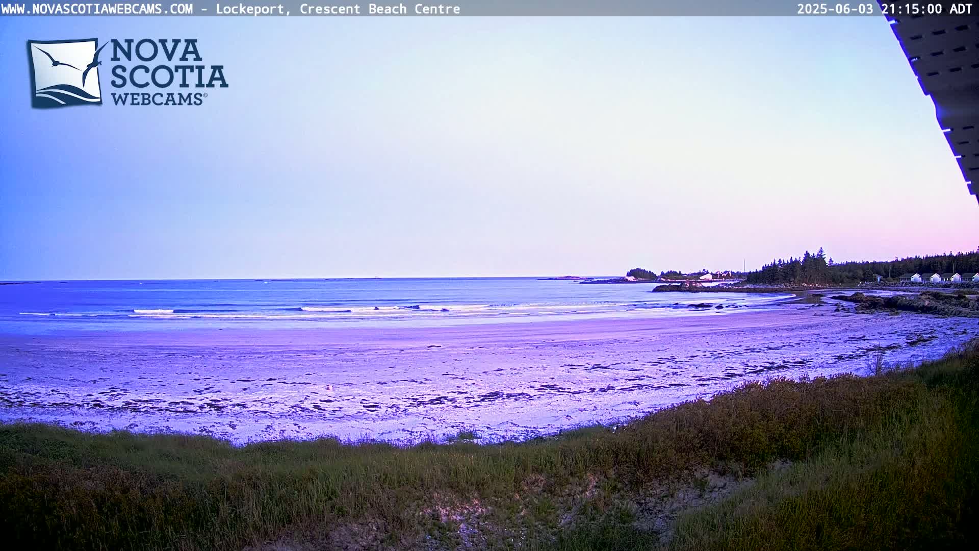 A sandy beach with gentle waves under a twilight sky is seen from behind a grassy dune.
