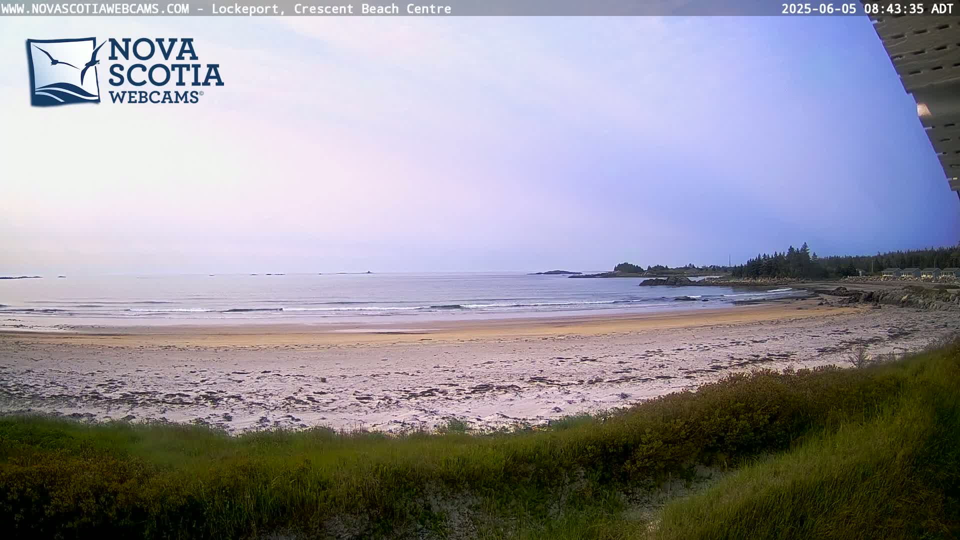 A sandy beach with small waves gently rolling onto the shore under a mostly clear, light-purple sky.