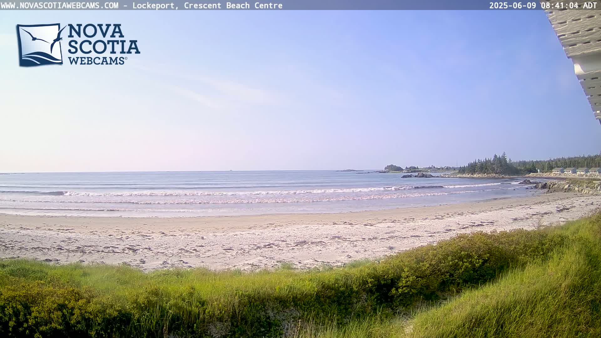 A sandy beach with small waves and a clear blue sky stretches toward a rocky shoreline and a calm ocean.