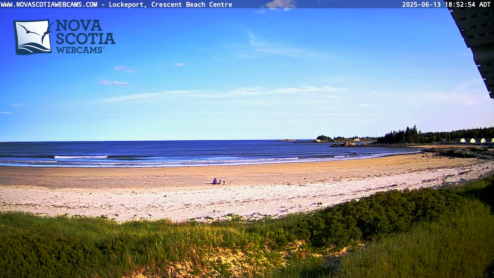 A sandy beach with a calm ocean under a clear blue sky, where a few people are sitting near the water's edge.