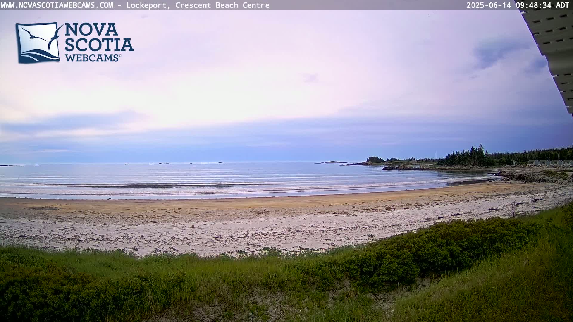 A sandy beach with gentle waves laps the shore under a mostly clear, pale sky, backed by low-lying land with dark green vegetation.