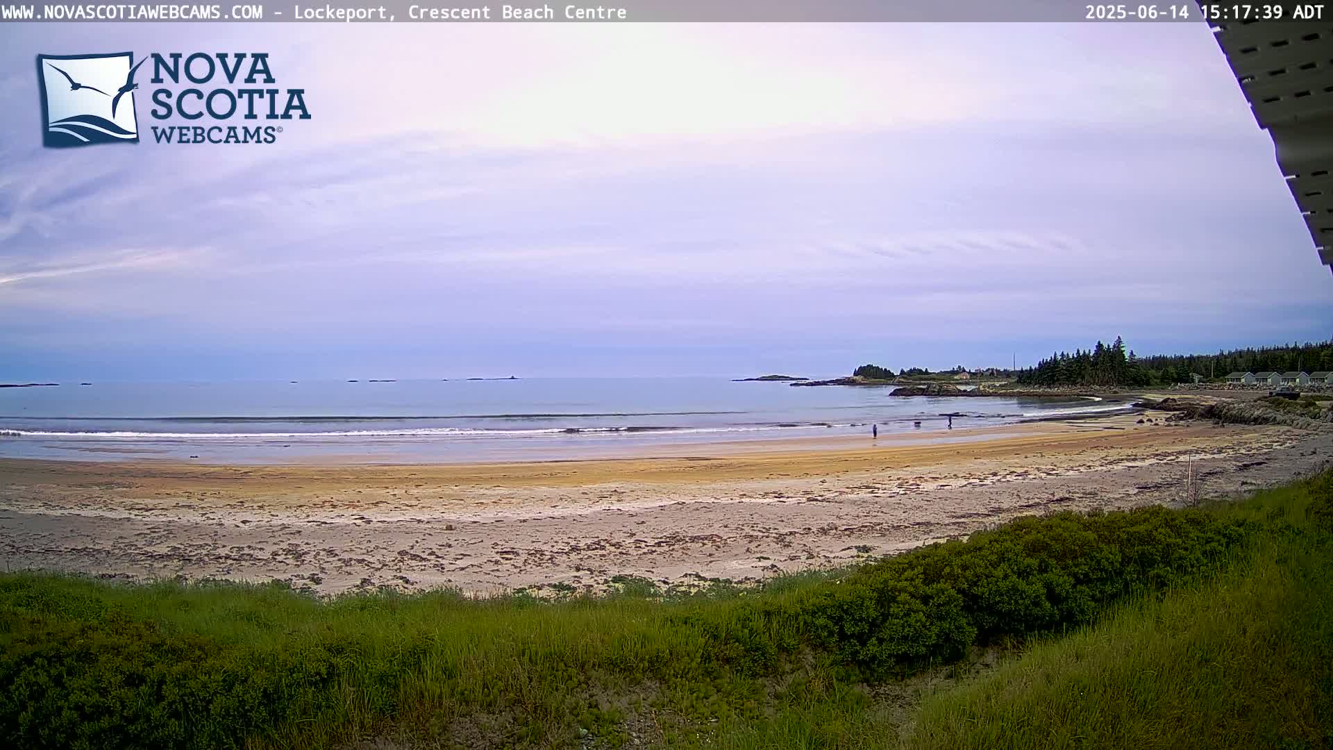 A sandy beach with calm ocean waves stretches out under a mostly cloudy sky, with a few people visible near the water's edge.