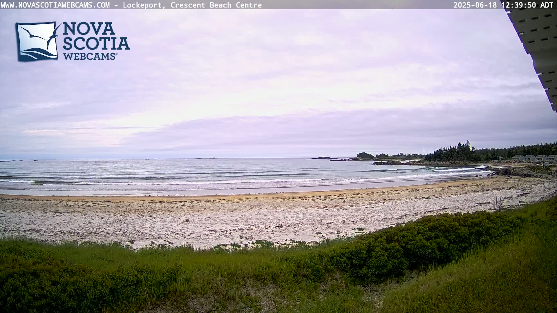 A sandy beach with gentle waves stretches to a calm ocean under a mostly cloudy sky.