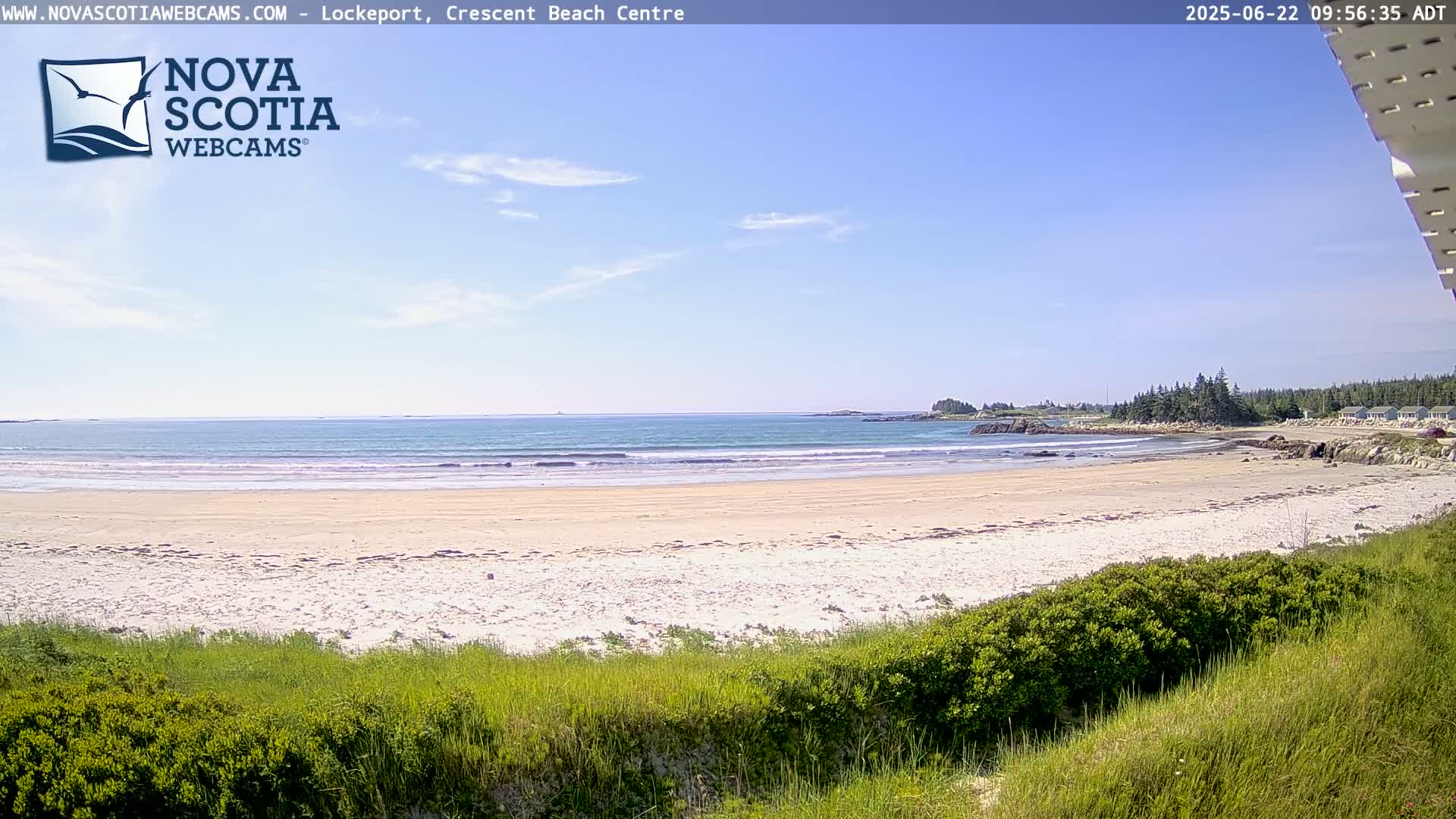A sandy beach with calm ocean waves stretches to a shoreline with trees and a few buildings under a mostly clear, sunny sky.