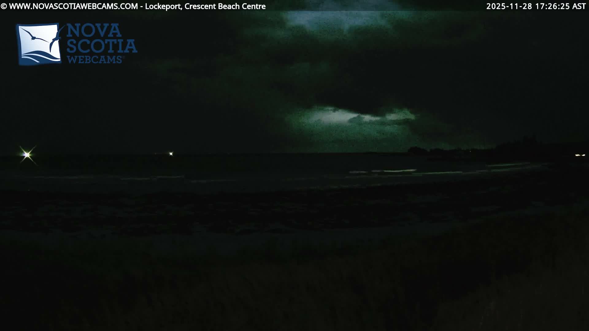 A dark, cloudy sky hangs over a dimly lit ocean with subtle waves and distant lights, indicating an overcast night.