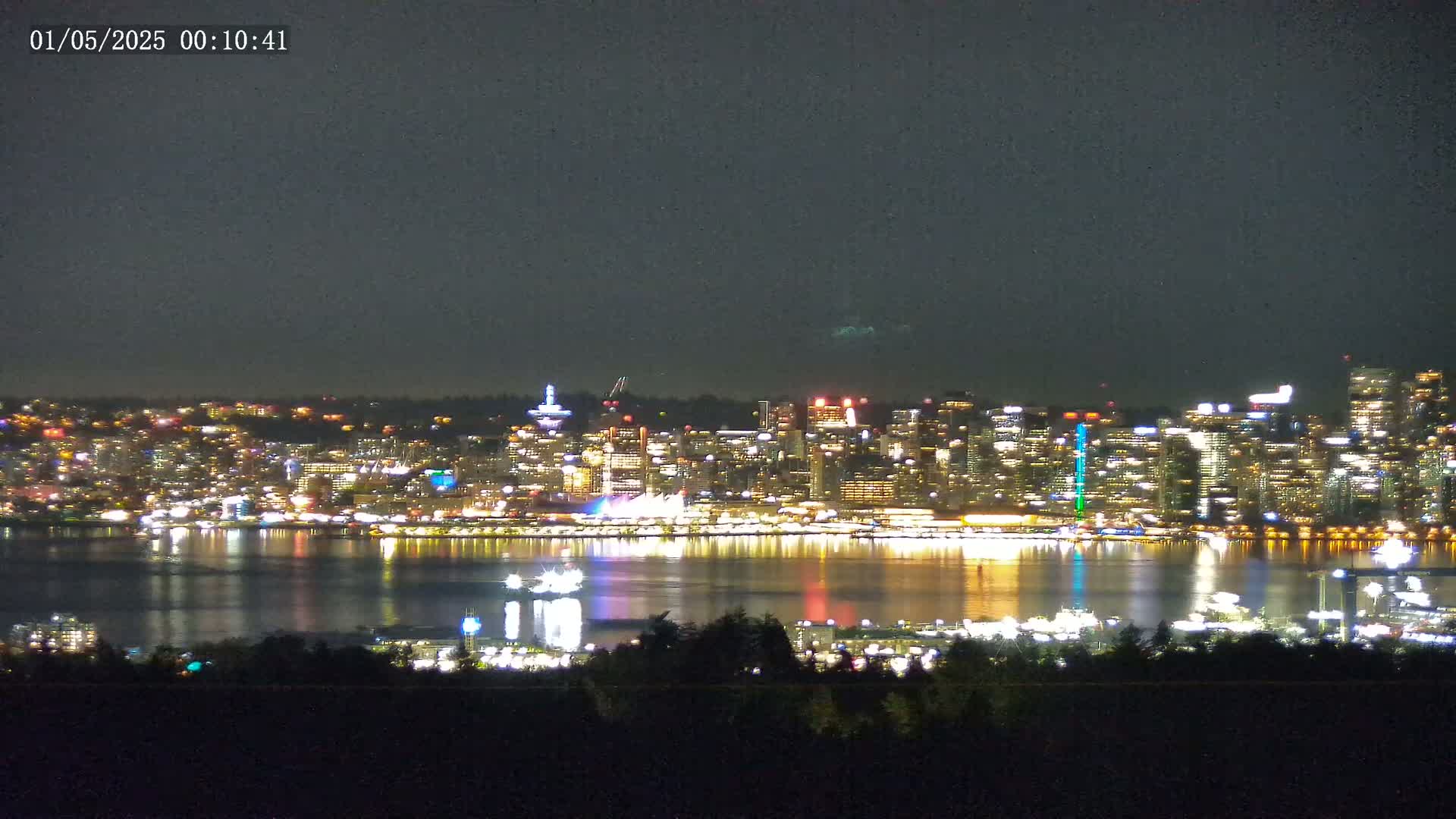 A brightly lit city skyline at night is reflected in a calm body of water, viewed from a distance across a dark expanse of land under a mostly clear night sky.
