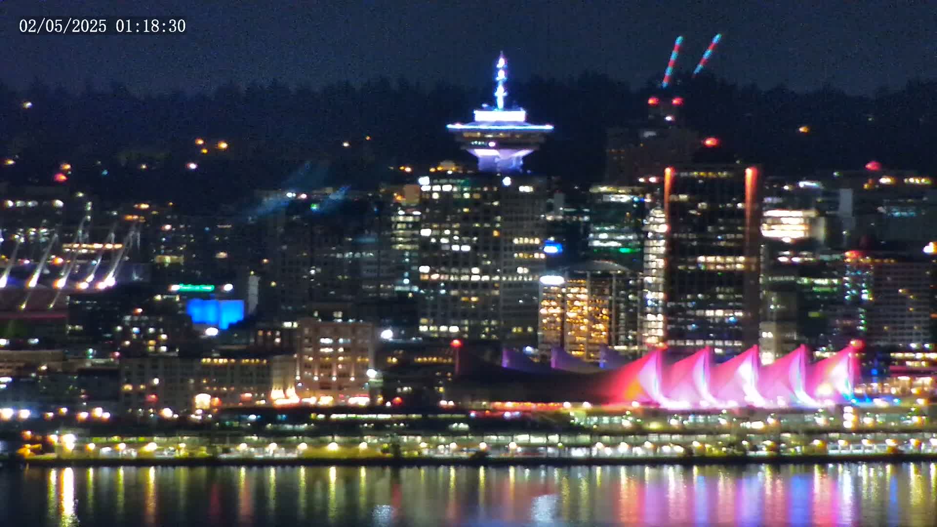 A brightly lit city skyline at night, reflected in calm water, features a prominent tower and illuminated structures along the waterfront under a clear, dark sky.