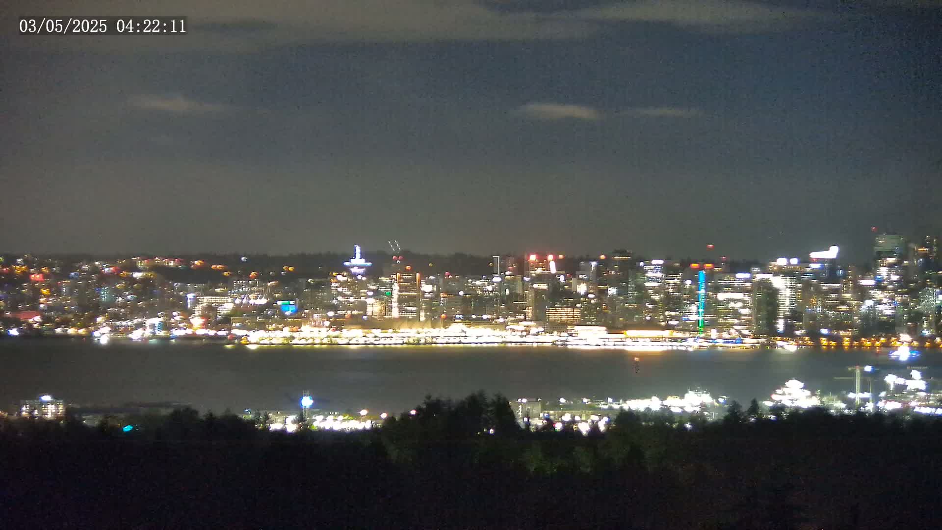 A nighttime view of a brightly lit city skyline across a dark body of water, under a partly cloudy sky.