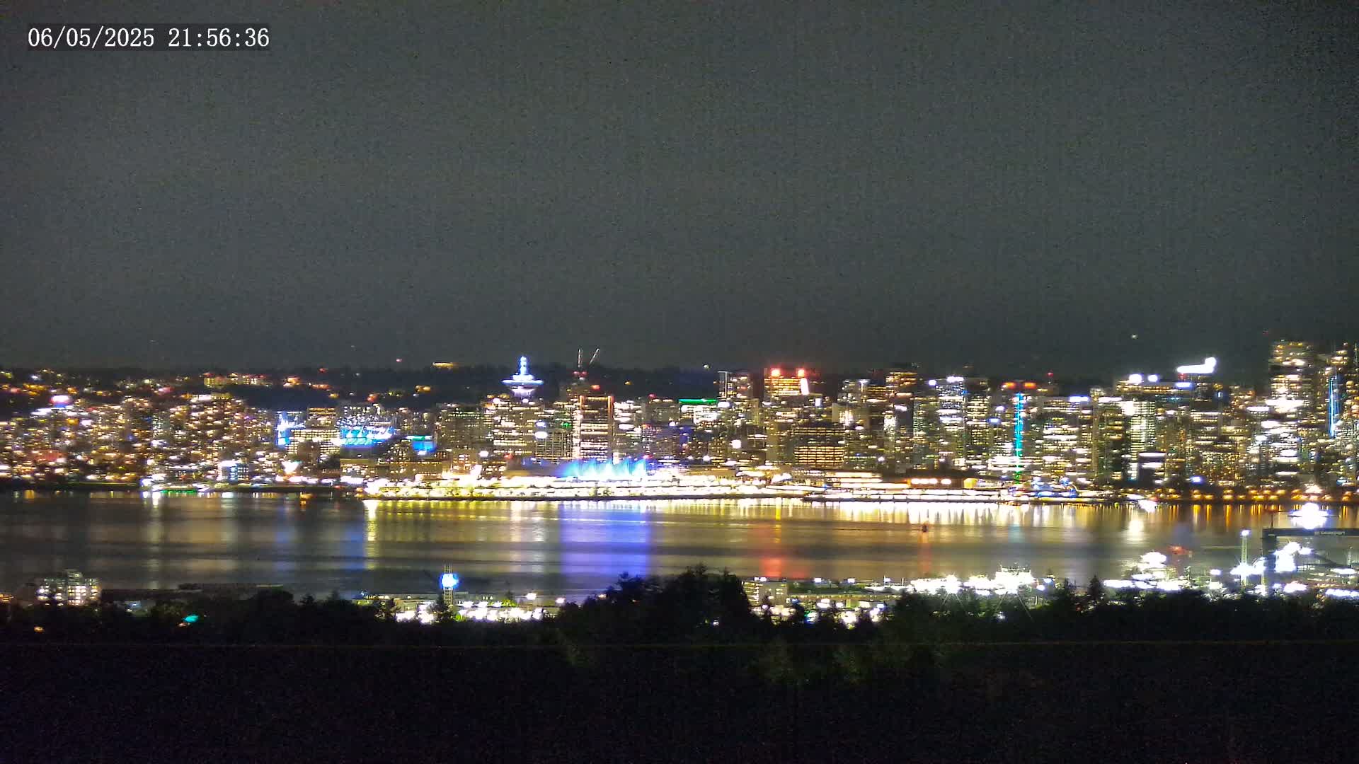 A brightly lit city skyline at night is reflected in a calm body of water, viewed from a distance under a dark sky.